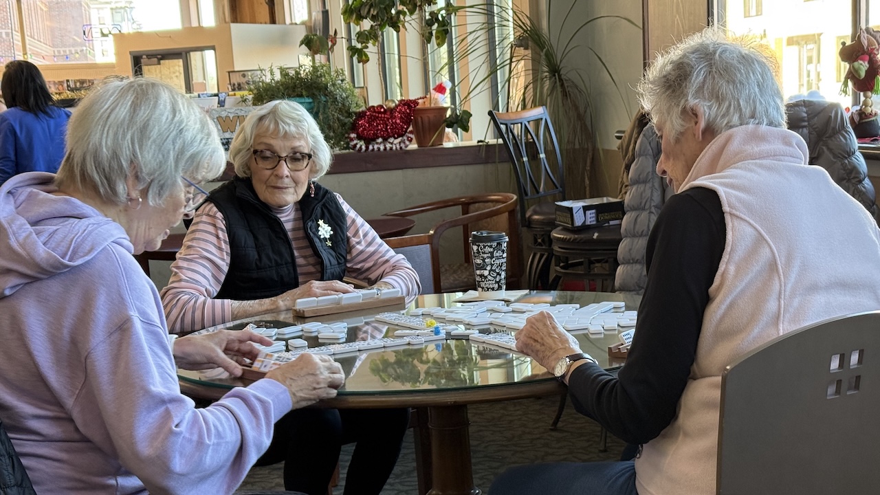 Three elderly women sitting around a glass table playing a game of Mexican Train Dominoes in a cozy café or community space, with festive holiday decorations in the background.