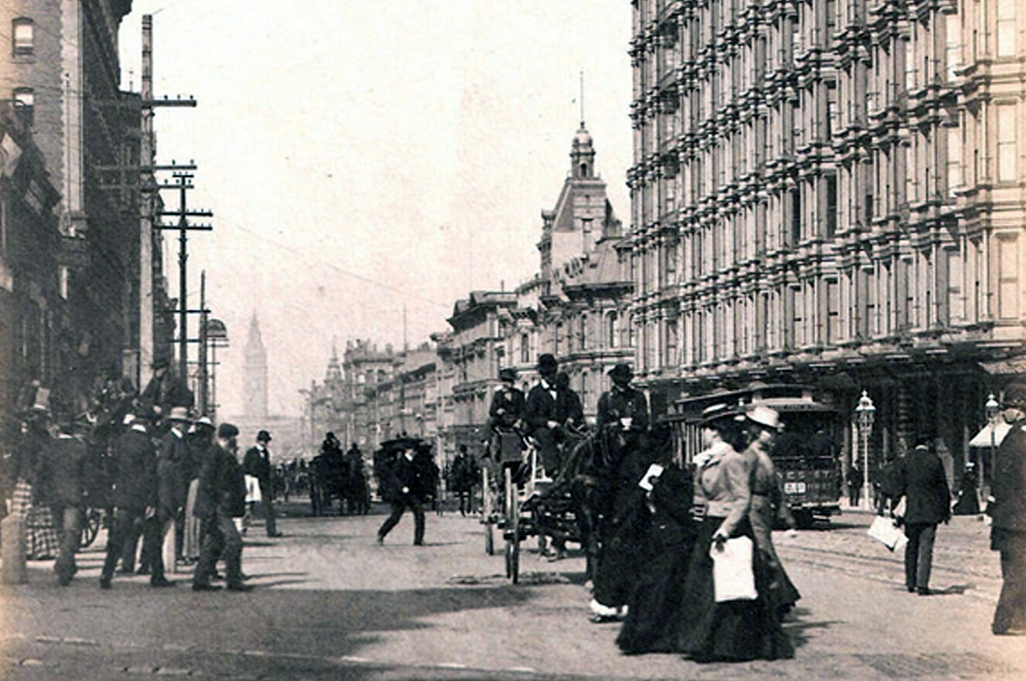 Historic black-and-white photo of a busy early 1900s city street with pedestrians, horse-drawn carriages, and ornate buildings, likely in San Francisco.